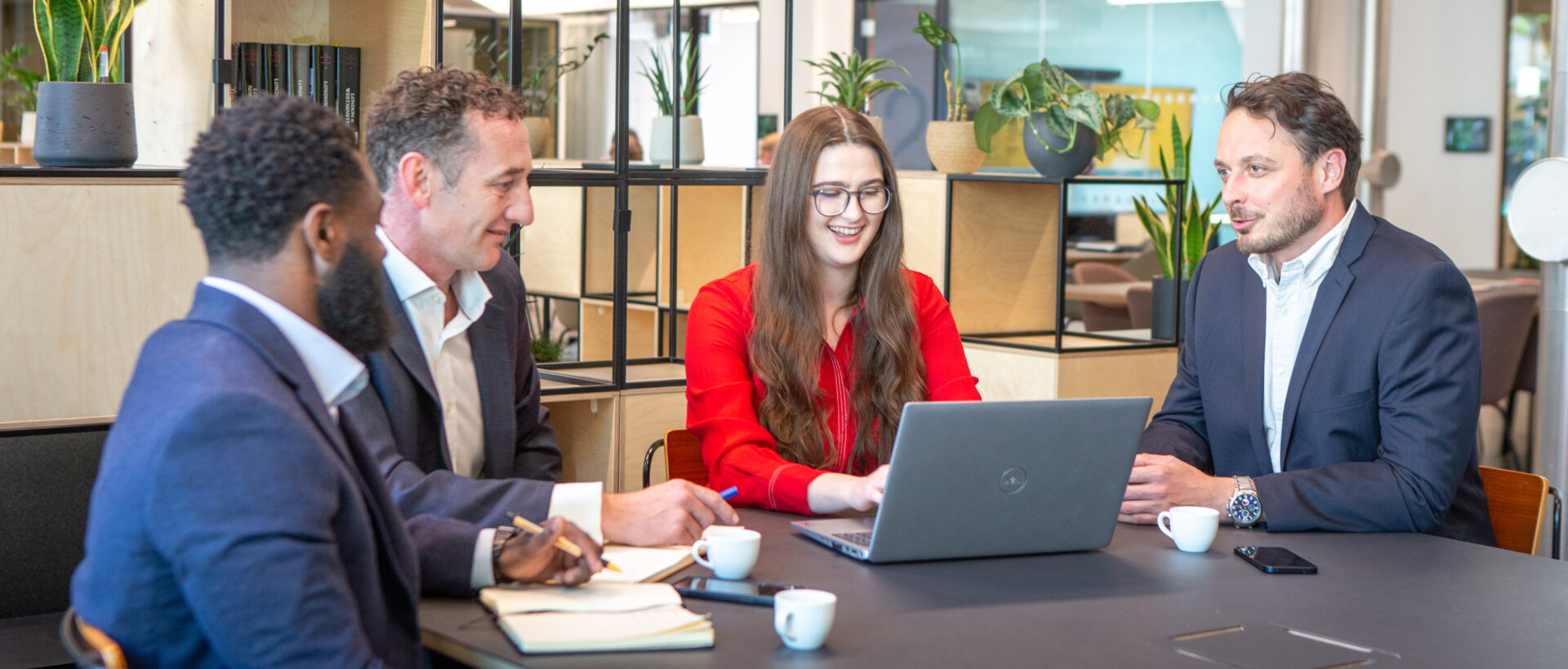 A woman in a red dress wearing glasses seated at a meeting table with an open laptop, with two men seated to her right and one an seated to her left, all looking at the laptop screen.