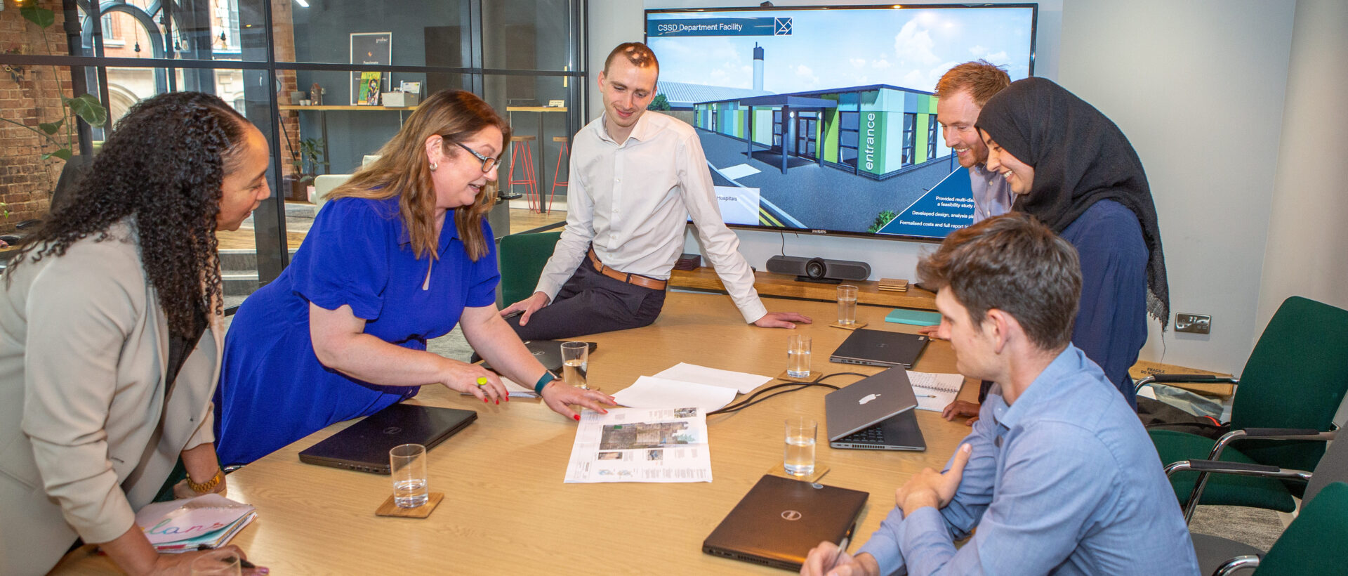 One man sitting on and two women leaning over a boardroom table showing some plans to one seated man, one standing man and one standing woman on the opposite side of the table.