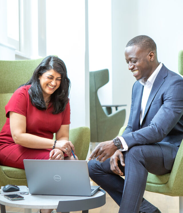 Woman in a red dress sat on a green chair next to a man in a blue suit also sat on a green chair, both are looking at an open laptop on the coffee table in front of them.