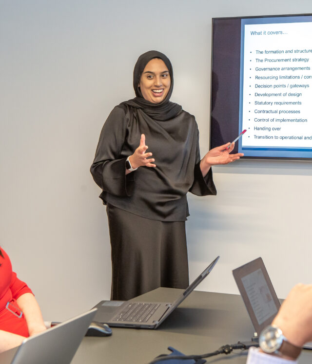 A woman wearing a hijab standing in front of a wall mounted screen presenting in to a seat woman and a seated man.