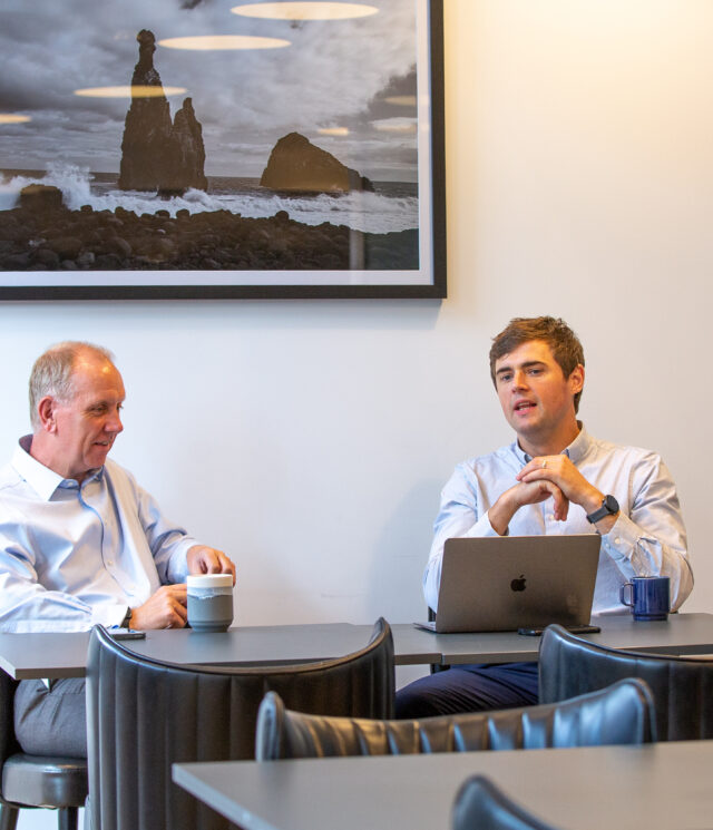 Two men sat at a table in an office breakout space with coffee and an open laptop having a conversation.