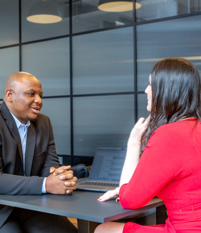 Woman in a red dress seated with her back to the camera having a conversation with a man in a grey suit seated in front of her, whilst a man in the background is working on his laptop.