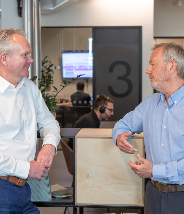 A man in a white shirt and blue trousers leaning on a freestanding shelving unit talking to a man in a light blue shirt and grey trousers, also leaning on the unit.