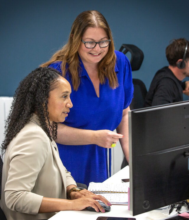 A woman sat at her desk using her mouse with a smiling woman stood next to her.