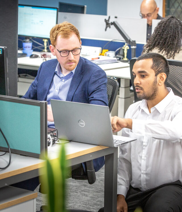 A man sat at his desk with a man crouching next to him pointing to his laptop screen on the desk.