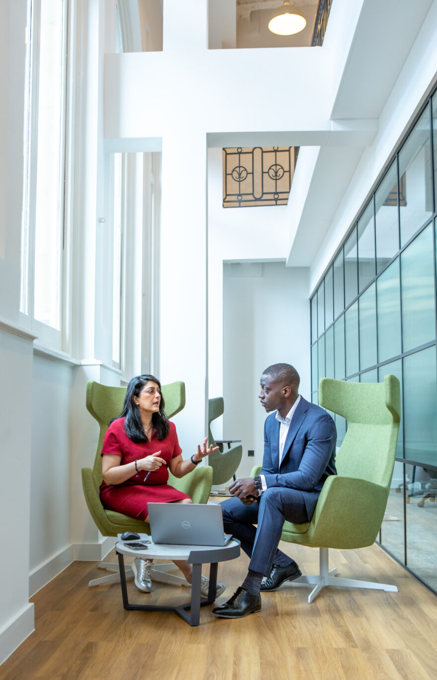 Woman in a red dress sat on a green chair next to a man in a blue suit also sat on a green chair, having a conversation over an open laptop on the coffee table in front of them.