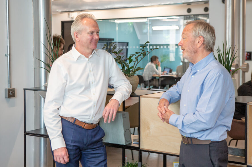 A man in a white shirt and blue trousers leaning on a freestanding shelving unit talking to a man in a light blue shirt and grey trousers, also leaning on the unit.