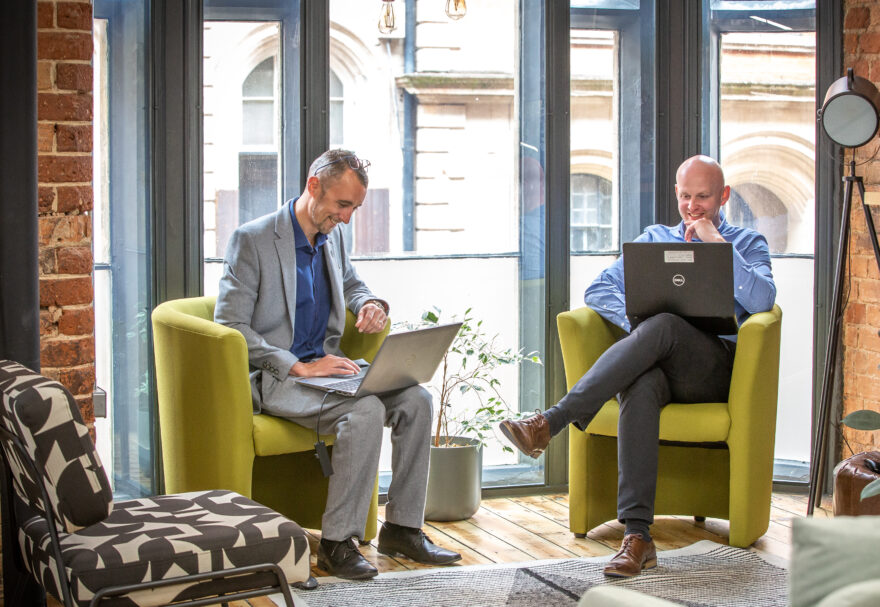 Two men sat in green chairs in a feature floor to ceiling window with laptops on their knees.