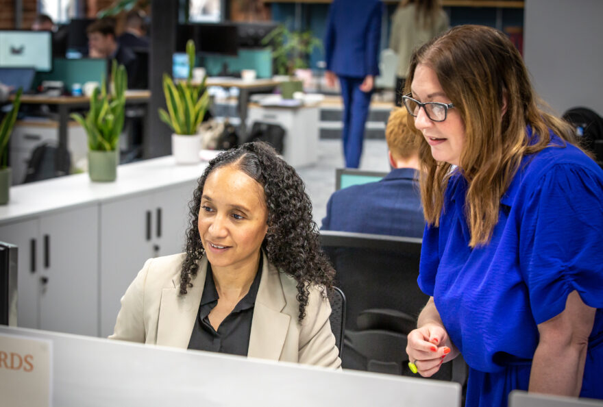 A seated woman looking at her monitor with a woman standing next to her also looking at the screen.