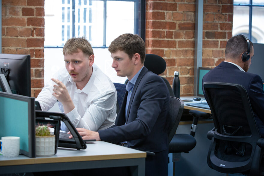 Two men sat at a desk discussing something one of them is pointing at on the screen, another man with his back to them on a video call.