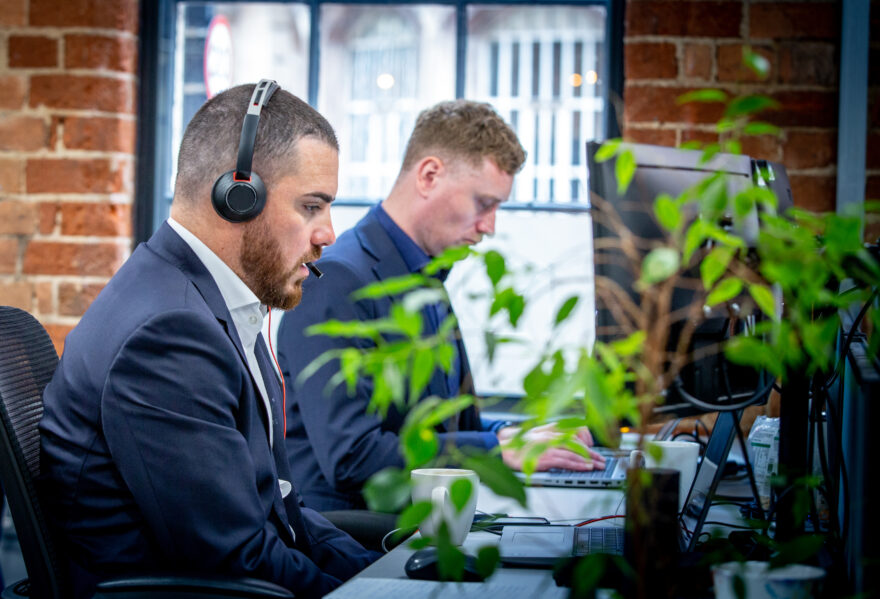 Two men sat at desks next to each other, the man in the foreground is wearing a headset and is on a video call.