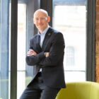 Man in dark grey suit perched on a green chair in a feature window with a brick wall and floor lamp in the background.