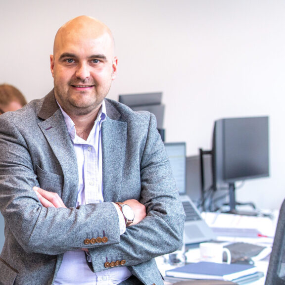 Man with folded arms sitting on the edge of his desk with colleagues working at desks behind him.