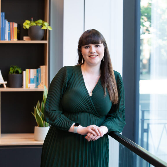 Woman in green dress leaning against a glass wall with shelves behind her.