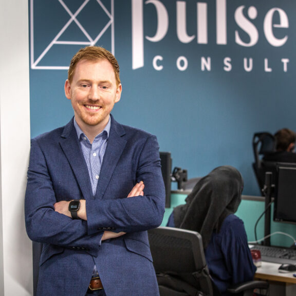 Man in a blue suit and folded arms leaning against a wall with colleagues working at desks behind him.