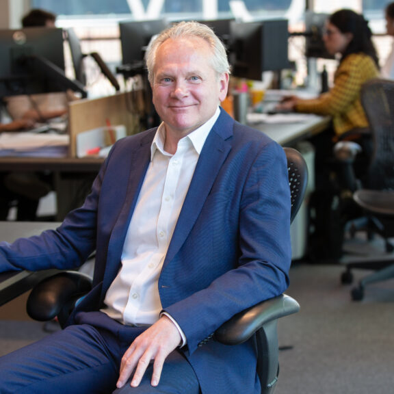 Man in blue suit sat at his desk with colleagues working at their desks in the background.