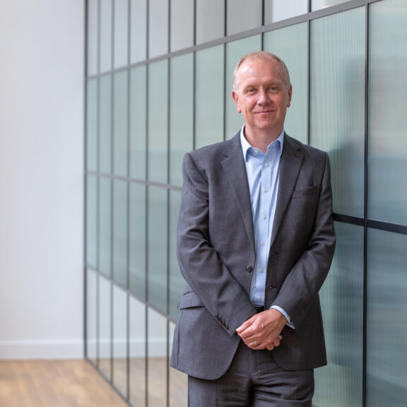 Man in grey suit with hands clasped leaning against a glass partition wall.