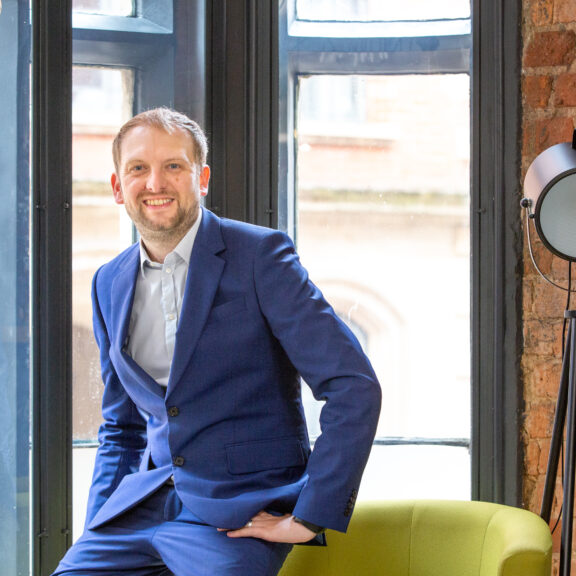 Man in blue suit perched on a green chair in a feature window with a brick wall and floor lamp in the background.