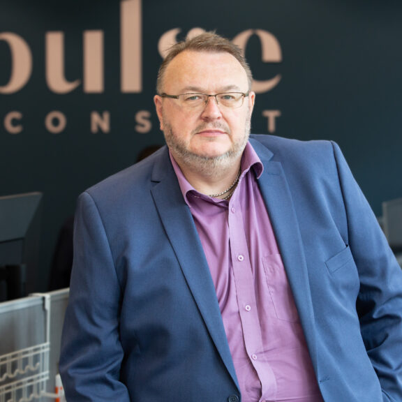 Man with glasses in a blue suit perched on the edge of his desk with colleagues working at their desks in the background.