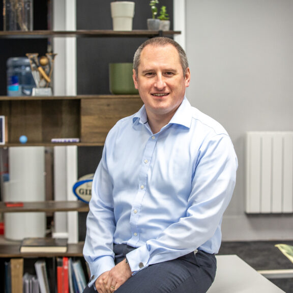 Man in a light blue shirt sat on a low cabinet with feature shelving behind him.