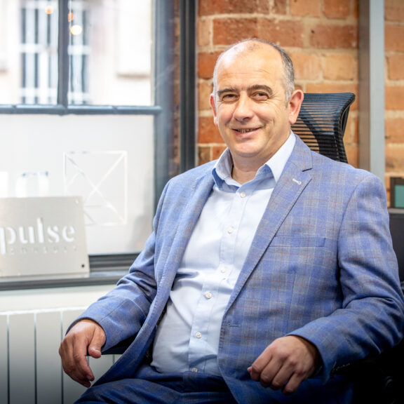 Man in a blue suit sat at his desk with a window and brick wall behind him.