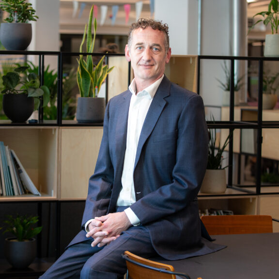 Man in a grey suit sat on a table in front of some feature shelving.