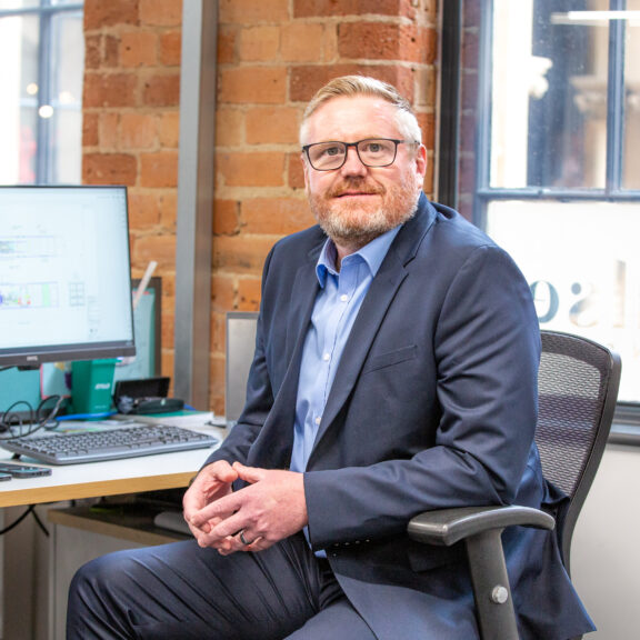 Man in a grey suit sat at his desk with windows and exposed brick behind him.